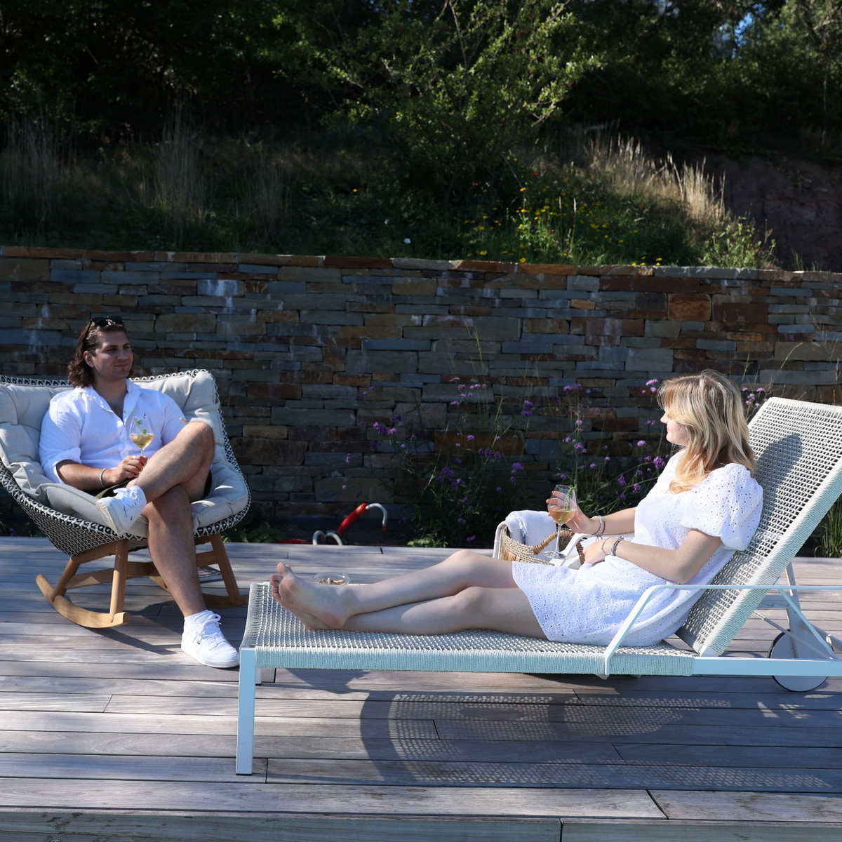 Gartenliege und Schaukel-Loungesessel auf einer Holzterrasse mit zwei entspannt sitzenden Personen.