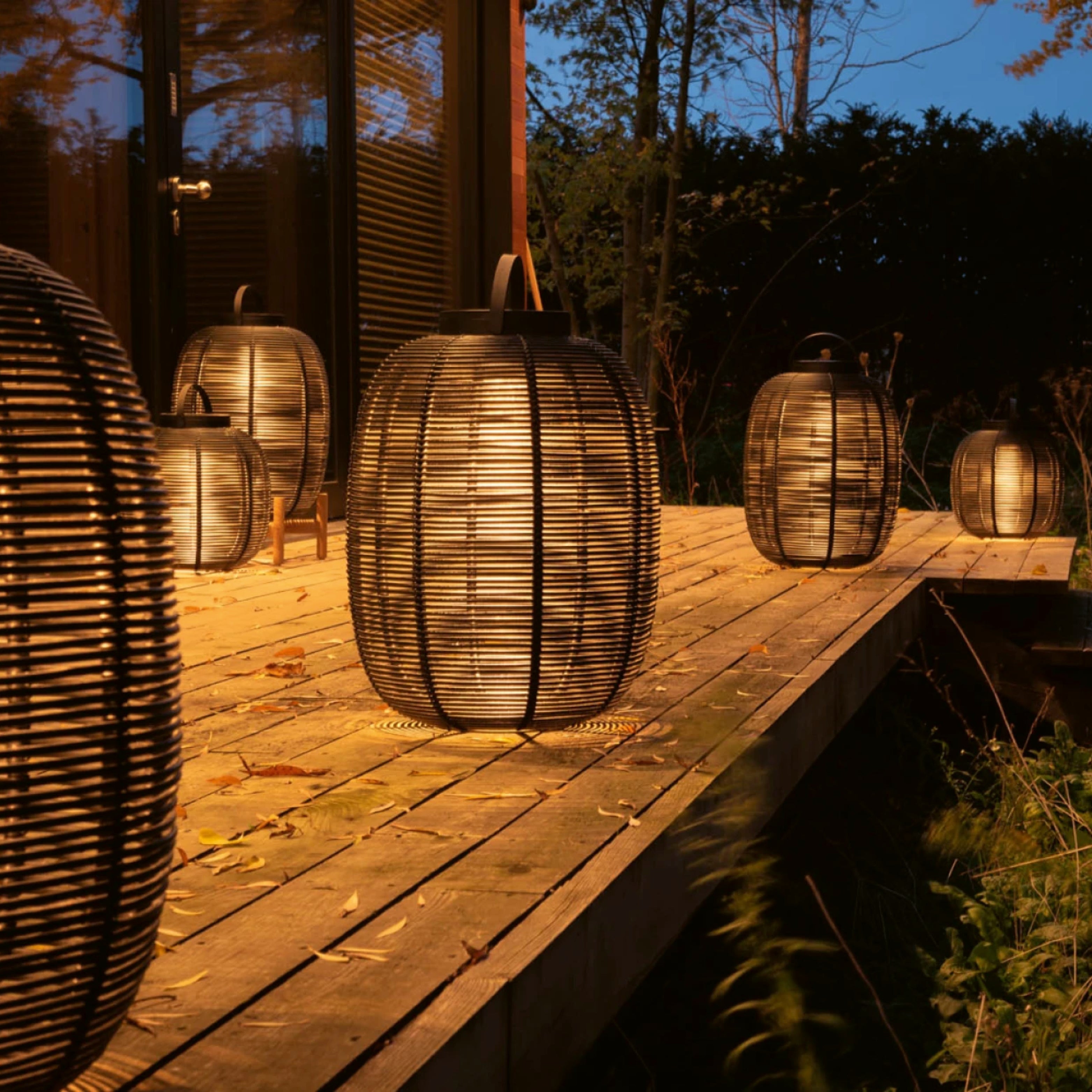 Mehrere schwarze Tika Lanterns von Vincent Sheppard leuchten warm auf einem Holzsteg im Garten bei Nacht – stimmungsvolle Gartenlampen-Szene.