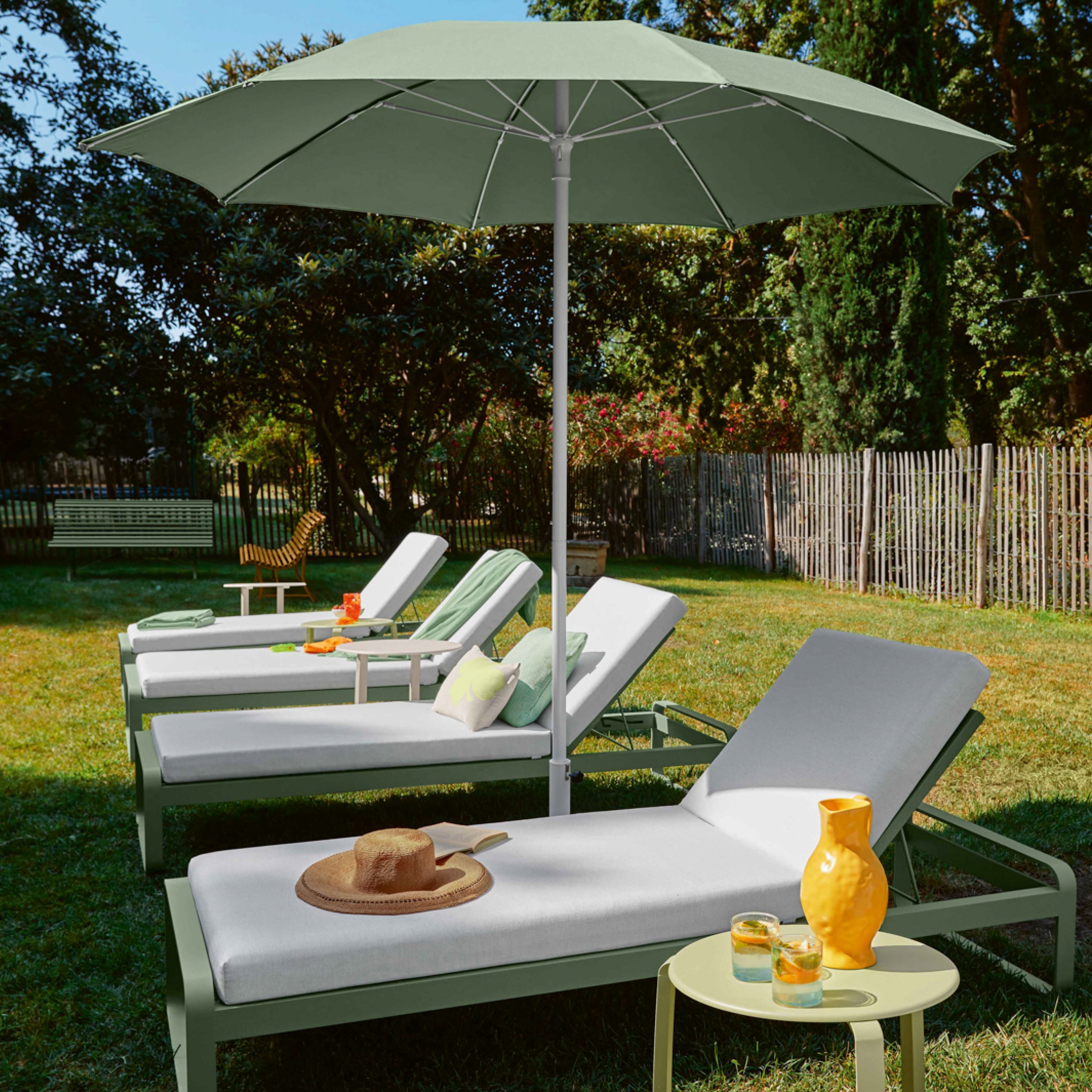 Bequeme Gartenliegen mit grauen Polstern unter einem grünen Sonnenschirm auf einer Wiese – Outdoor-Entspannung mit Drinks und Snacks