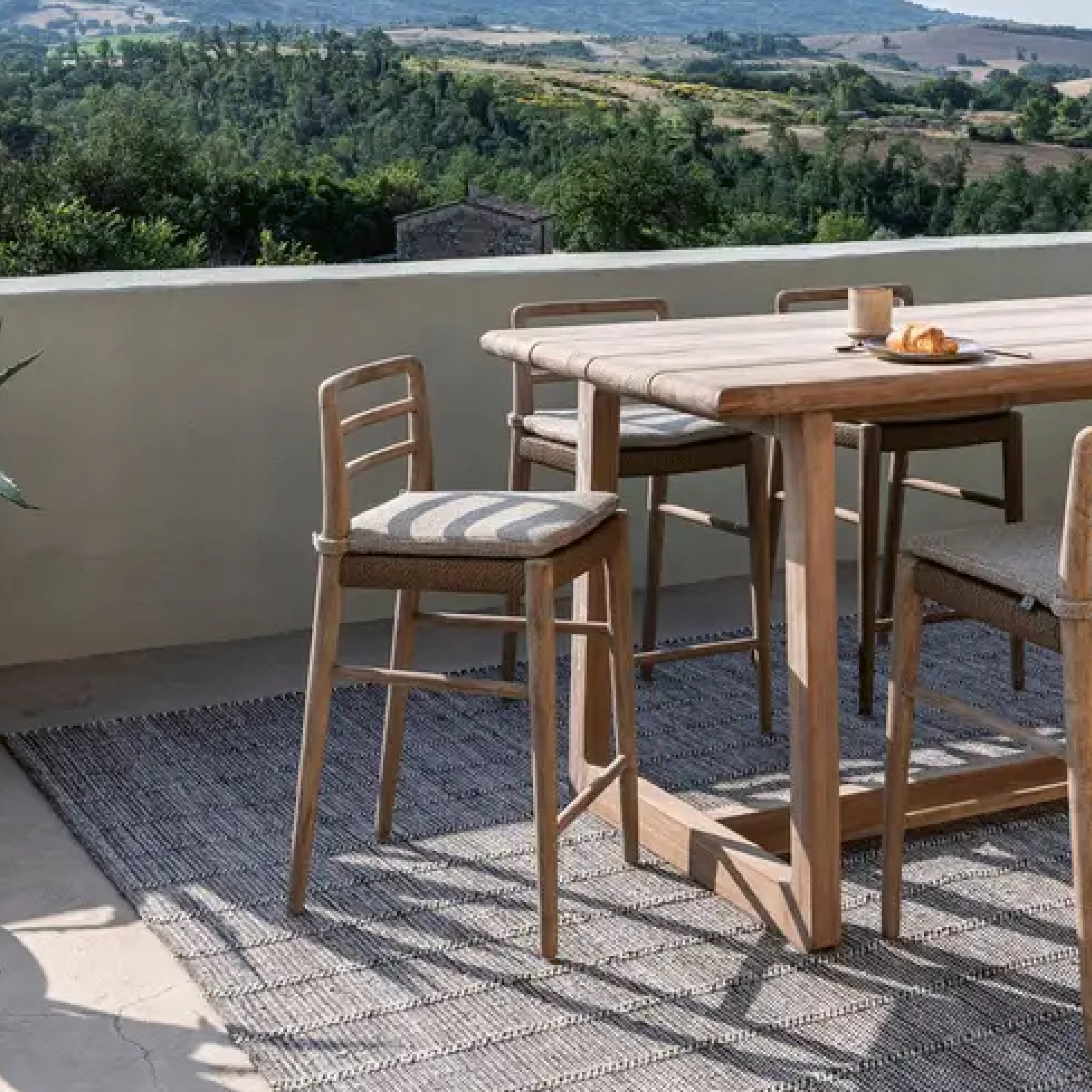 Hochwertiger Bartisch mit passenden Barstühlen von Gommaire auf einer sonnigen Terrasse mit Ausblick ins Grüne – minimalistisches Outdoor-Design in Naturtönen.