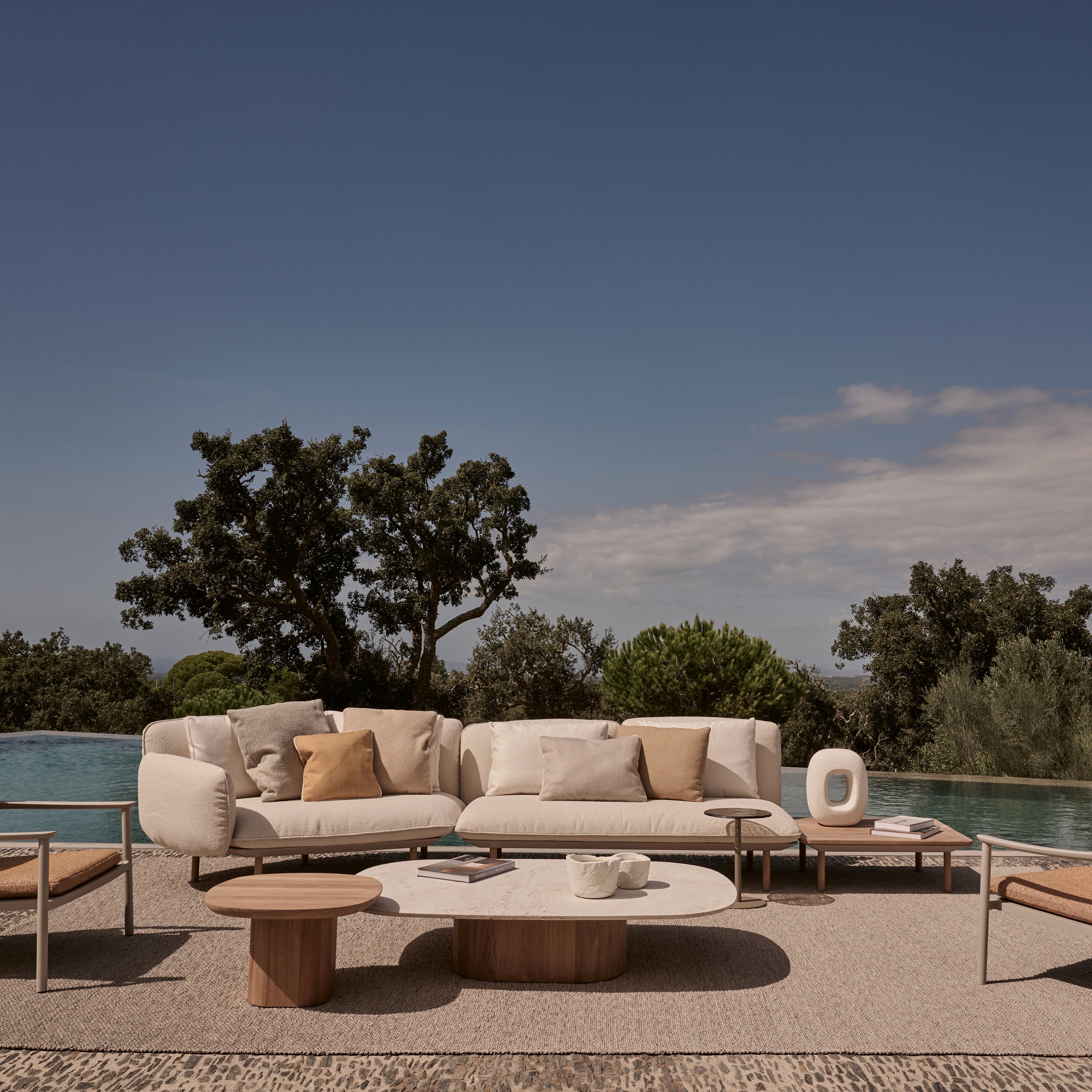 Elegante Outdoor-Lounge mit hellem Sofa, Sesseln und Couchtischen vor einem Pool, umgeben von mediterraner Landschaft – Holiday Homestorys Urlaubsfeeling im Garten.