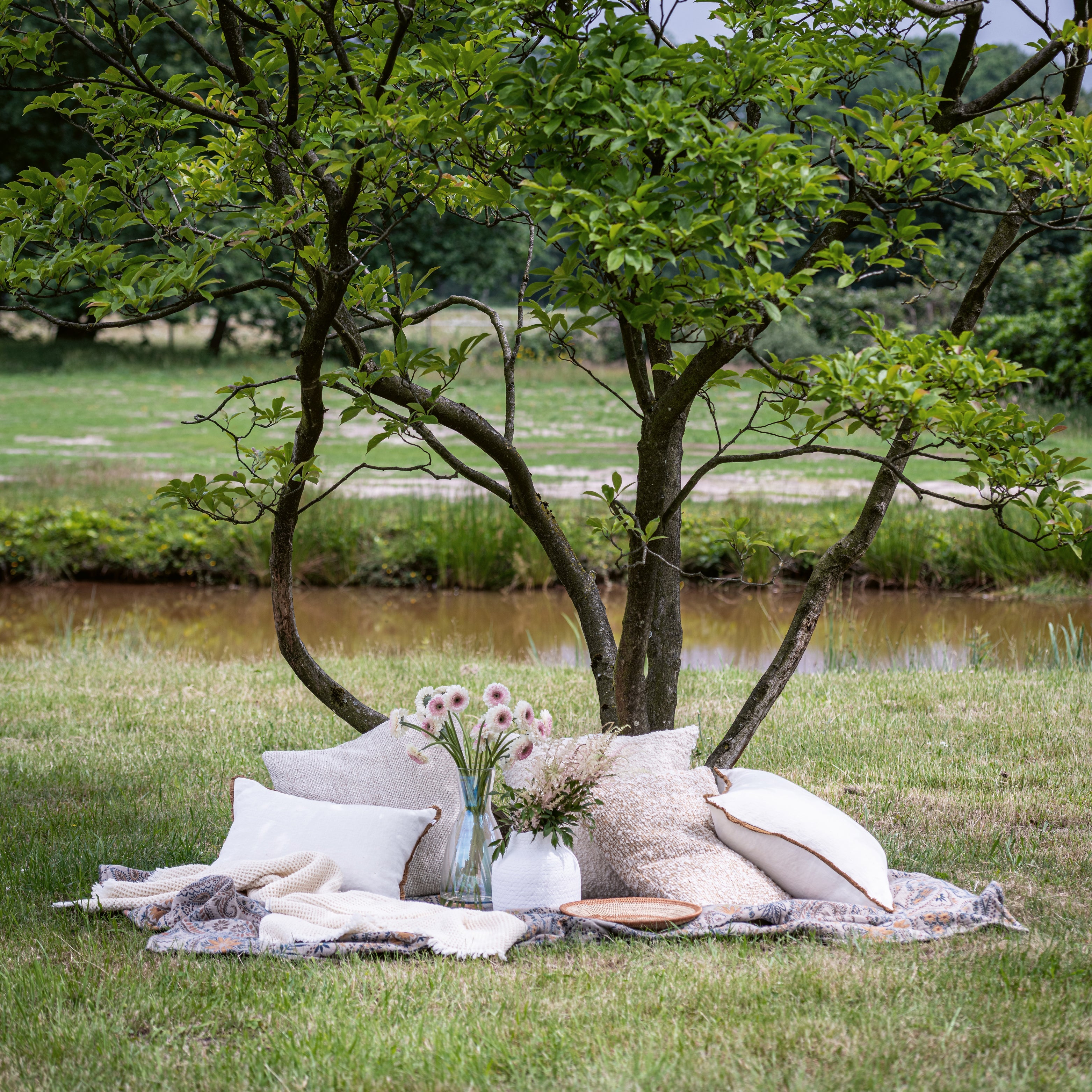 Dekorationskissen und Leinenkissen in Naturtönen auf einer Decke im Gras unter einem Baum, kombiniert mit Vasen und Blüten – gemütliche, natürliche Kissendeko.