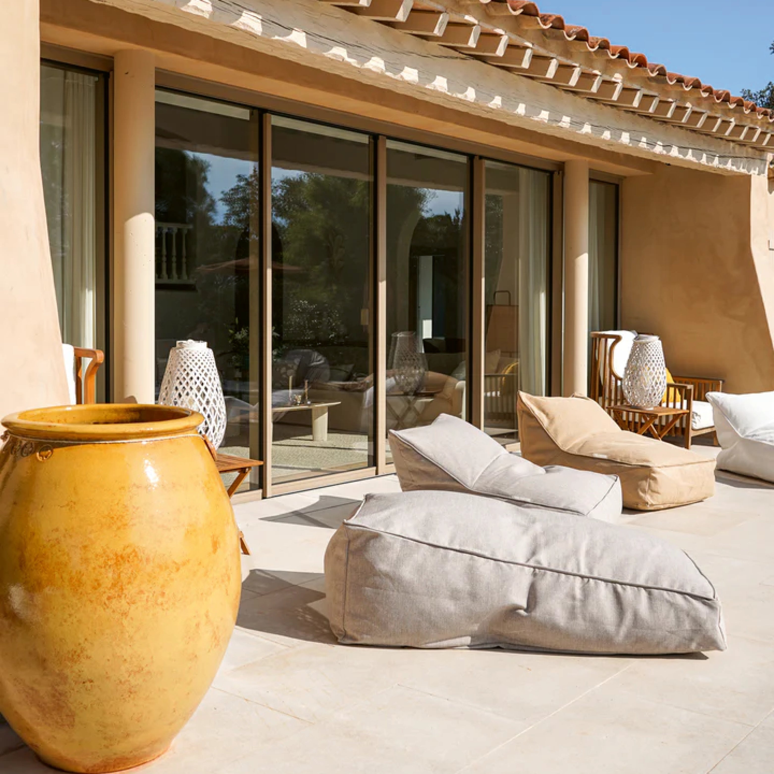Moderne Outdoor Sitzsäcke in Naturtönen auf sonniger Terrasse vor einem Haus mit bodentiefen Fenstern und mediterranem Flair.