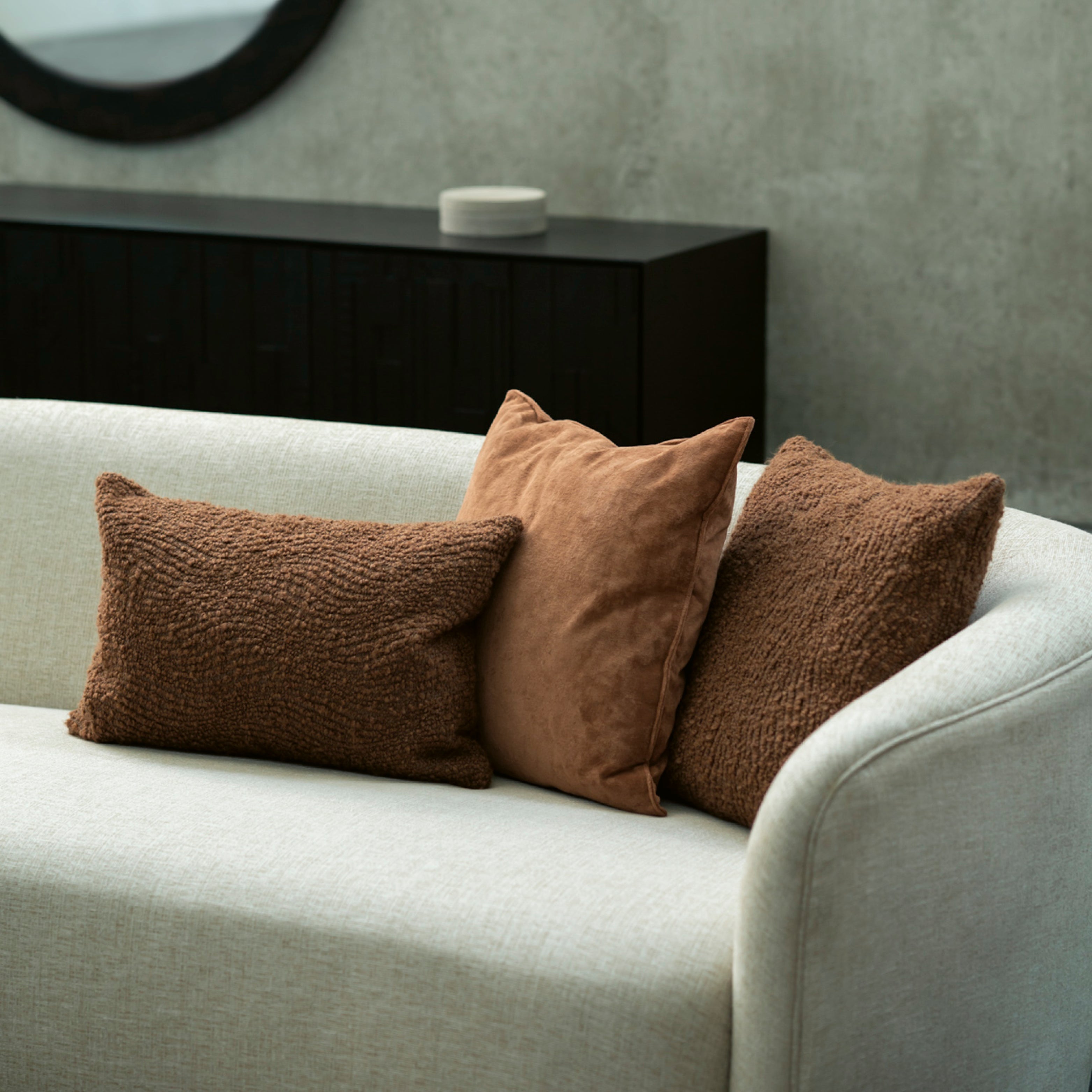 Drei Zierkissen aus Textil auf hellem Sofa vor dunklem Sideboard