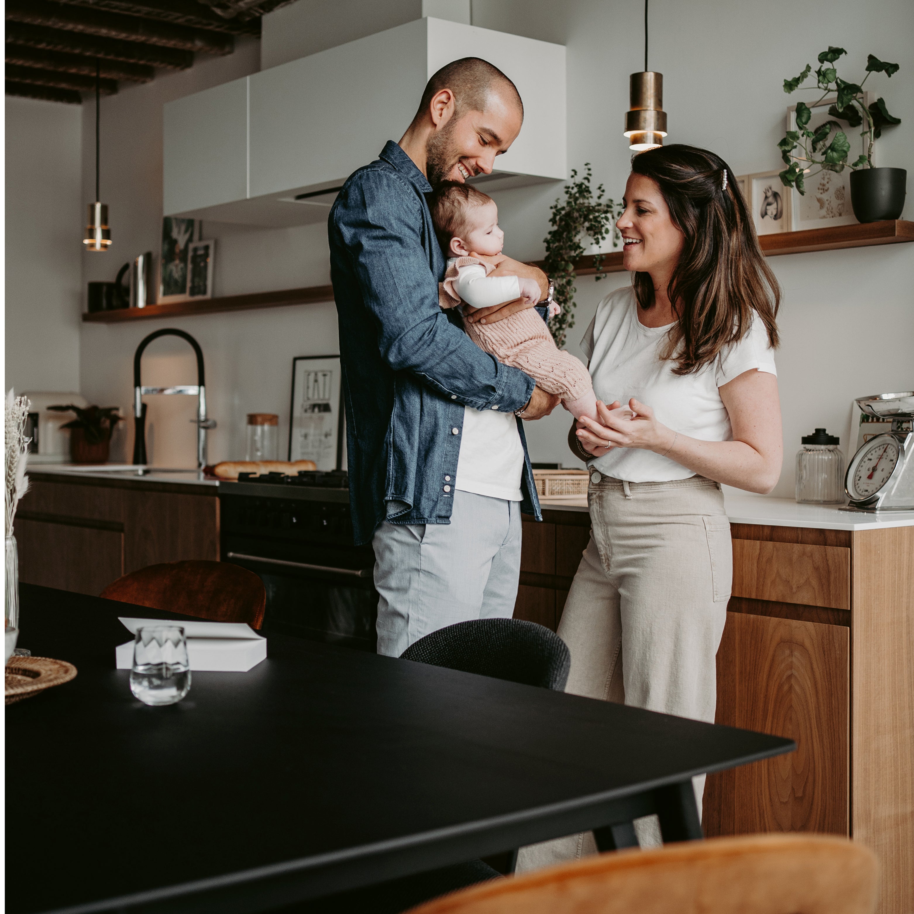 Junge Familie mit Baby in der Küche, Eltern halten lächelnd ihr Kind im Arm, rechts zwei Kinder spielend unter einer Decken-Höhle – warmes, liebevolles Familienzuhause.