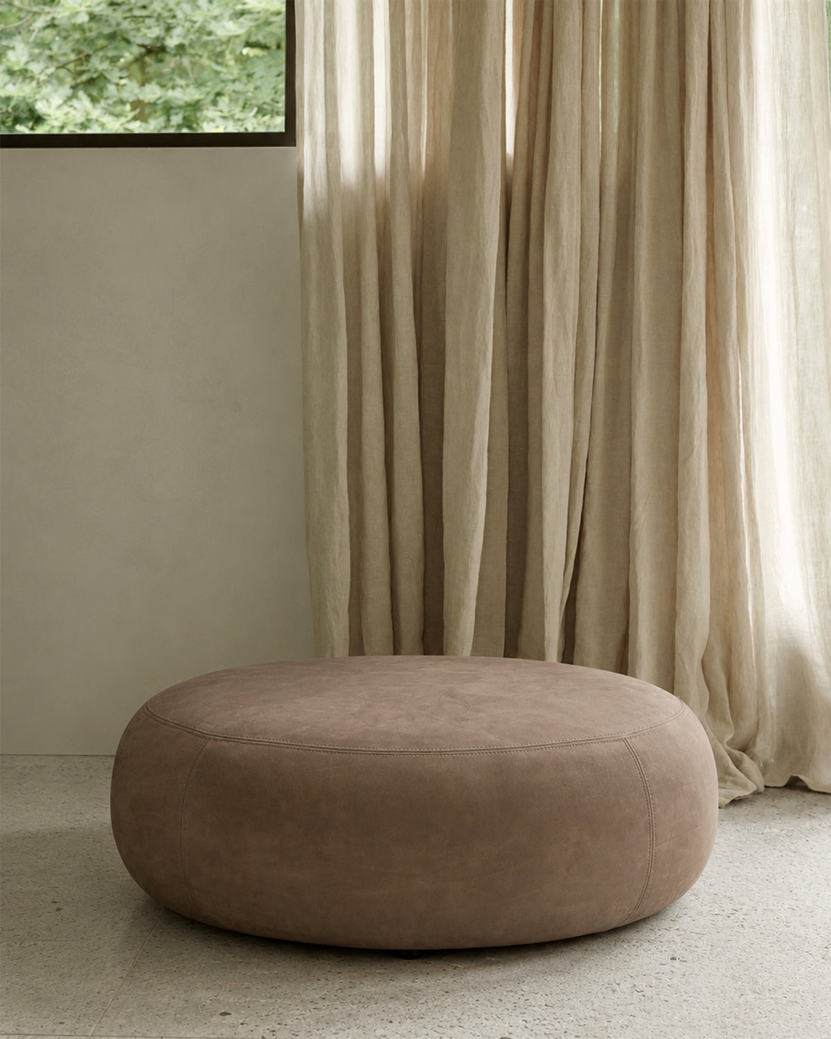 Ethnicraft ORB ottoman in fawn nubuck leather, used as a padded footstool in front of a lounge chair in a cozy living room.