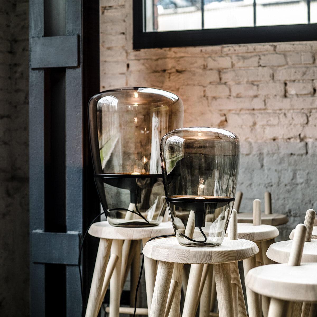 Zwei BALLOONS PC858 Lampen von Brokis – eine mit Rauchbraunem Glas und Kupferreflektor, eine mit Rauchgrauem Glas und Messing – auf Hockern vor Klinkerwand